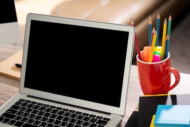 Offener Laptop mit schwarzem Bildschirm neben rotem Becher mit bunten Buntstiften auf einem Schreibtisch