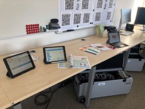 Foto aus der Medienwerkstatt in Kronshagen. Unter dem Arbeitsplätzen befinden sich Koffer mit iPads. Auf den Tischen stehen iPads und Unterrichtsmaterialien,