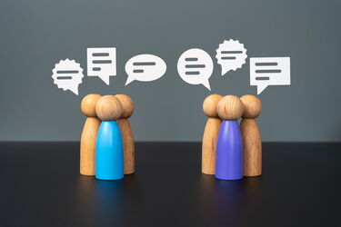 Zwei Gruppen aus je vier Holzfiguren, zwei Figuren in der Mitte sind blau und lila, darüber Sprechblasen als Symbol für Kommunikation.