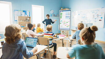 Ein Lehrer steht an einem großen Display vor einer Klasse, während die Schüler aktiv zuhören