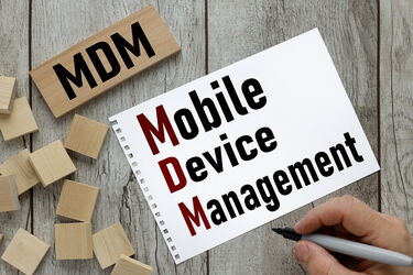 Hand schreibt auf einem Notizblock die Wörter 'Mobile Device Management' mit hervorgehobenen roten Anfangsbuchstaben M, D, M auf einem Holztisch mit Holzklötzen und einem Schild mit 'MDM'