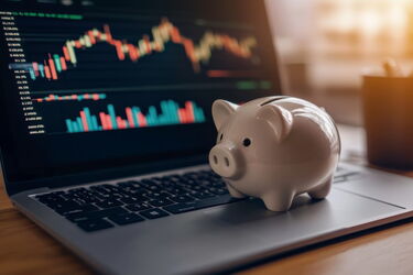 Weißes Sparschwein auf Laptop mit Finanzdiagrammen und Kursverläufen auf dem Bildschirm.
