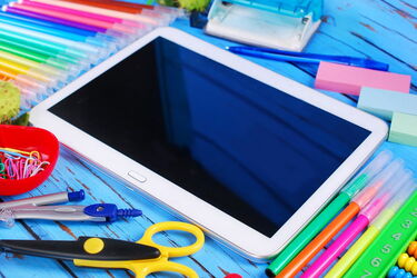Tablet auf Tisch umgeben von bunten Stiften, Schere, Zirkel und Büroklammern.